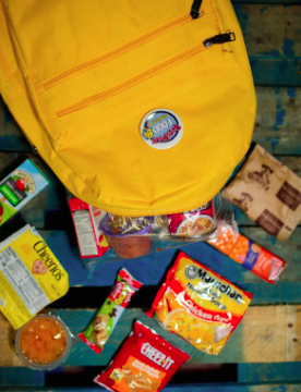 A yellow backpack on a picnic table surrounded by snacks and shelf-stable food for the weekend backpack program